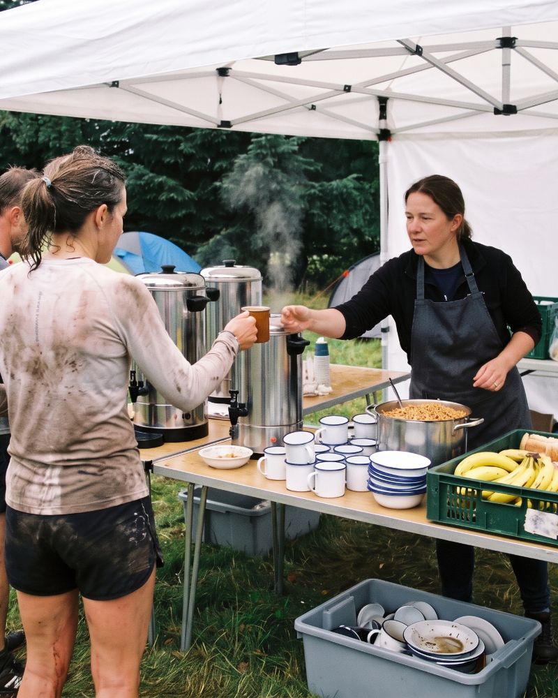 Helferin reicht einer verschlammten Läuferin einen Becher über den Tisch im Küchenzelt